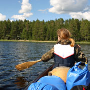 järvimaisema, jossa näkymä kanootista, edessä melojan selkä, taustalla metsäinen ranta.