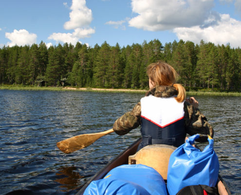 järvimaisema, jossa näkymä kanootista, edessä melojan selkä, taustalla metsäinen ranta.