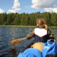 järvimaisema, jossa näkymä kanootista, edessä melojan selkä, taustalla metsäinen ranta.