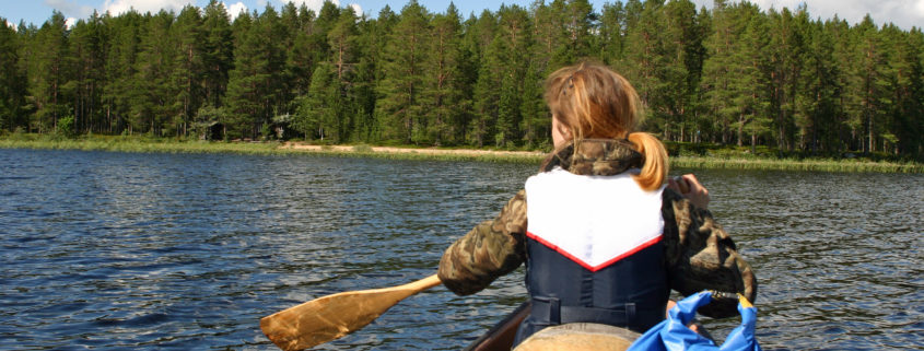 järvimaisema, jossa näkymä kanootista, edessä melojan selkä, taustalla metsäinen ranta.